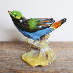 Op de afbeelding: Een kleurrijk keramisch vogelbeeldje. De vogel heeft een groene kop, een zwarte rug, een blauwe buik en oranje en rode accenten op de vleugels. Hij zit op een beige voet met een gele onderkant. Het beeldje staat tegen een neutrale achtergrond.