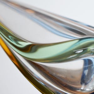 May include: Abstract glass sculpture with flowing lines and a blend of colors. The piece features shades of yellow, green, blue, and clear glass, creating a dynamic and artistic form. The design is smooth and curved.
