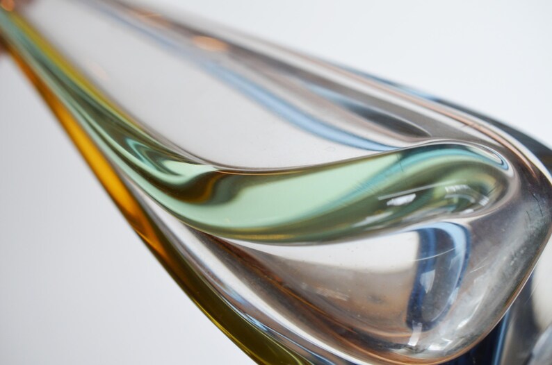 May include: Abstract glass sculpture with flowing lines and a blend of colors. The piece features shades of yellow, green, blue, and clear glass, creating a dynamic and artistic form. The design is smooth and curved.