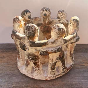 May include: A circular clay sculpture featuring six stylized figures standing in a ring with their arms linked around each other. The figures have a weathered, off-white finish with dark brown, earthy patches and textured, hand-molded details.