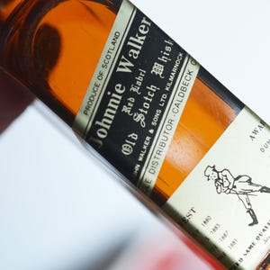 May include: A brown glass bottle of Johnnie Walker Red Label Old Scotch Whisky. The label is white with black text and a black and white illustration of a man walking. The bottle is labeled "Produce of Scotland" and "John Walker & Sons Ltd. Kilmarnock".