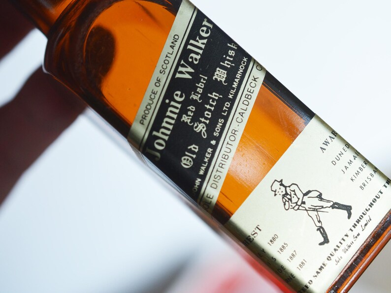 May include: A brown glass bottle of Johnnie Walker Red Label Old Scotch Whisky. The label is white with black text and a black and white illustration of a man walking. The bottle is labeled "Produce of Scotland" and "John Walker & Sons Ltd. Kilmarnock".