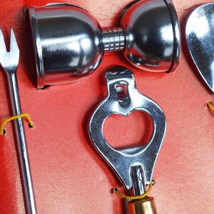 May include: A set of four silver bar tools, including a bottle opener, a cocktail fork, a jigger, and a spoon. The tools are arranged on a red background.