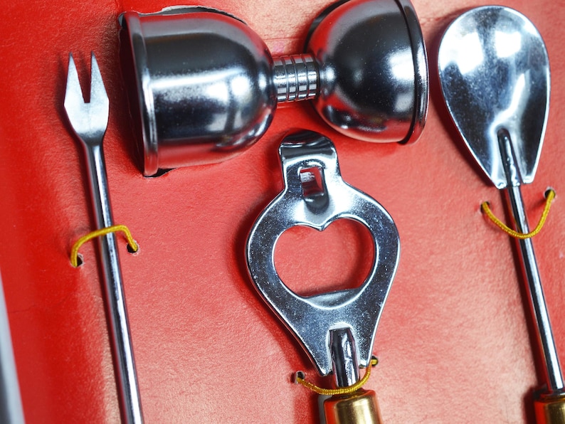 May include: A set of four silver bar tools, including a bottle opener, a cocktail fork, a jigger, and a spoon. The tools are arranged on a red background.