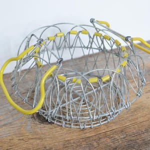 Op de afbeelding: Een zilveren draadmand met gele plastic accenten. De mand is inklapbaar en heeft een ronde vorm.