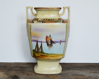 May include: A cream-coloured ceramic vase with gold accents and a painted scene of sailboats on a body of water. The vase has two handles and a square body.