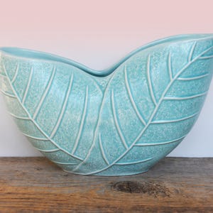 May include: A light blue ceramic vase shaped like two leaves. The vase has a textured surface and a glossy finish.