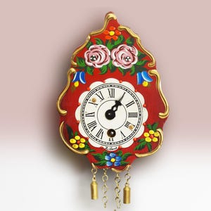 May include: A red wall clock with a scalloped shape and gold-painted edges. The clock face features a white background with black Roman numerals and black hands. It is decorated with hand-painted floral motifs in pink, yellow, and blue, and has three hanging gold-toned metal weights.