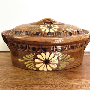 May include: Brown ceramic casserole dish with a lid, featuring a floral design in yellow and green. The dish has a handle on the lid and decorative detailing around the rim. A rustic, handmade kitchenware item.