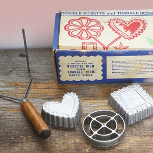 May include: A vintage set of metal molds for making rosette and timbale cookies. The set includes two heart-shaped molds, a square mold, and a round mold with a star design. The molds are packaged in a blue and white box with the text "Double Rosette and Timbale Iron" printed on the front.