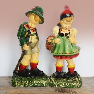 May include: Two ceramic figurines of children. One is a boy in a green hat, backpack, and shorts, holding a walking stick. The other is a girl in a red hat and green dress, holding a basket of flowers. Both stand on green bases.