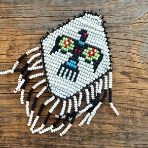 May include: A diamond-shaped beaded patch featuring a central black bird motif with white, red, green, and blue accents. The patch is bordered by a black beaded edge and features long, dangling fringe strands made of alternating black and white beads.