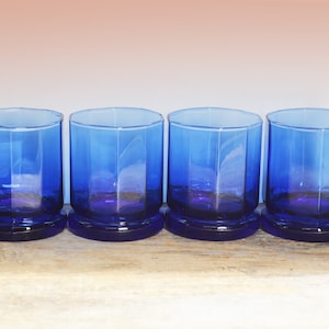 May include: Set of four cobalt blue glass tumblers with a faceted design. The glasses are sitting on a wooden surface.