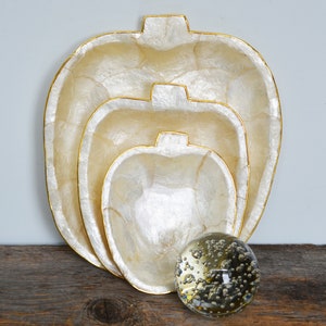 Può includere: Tre ciotole decorative bianche perlacee con bordo dorato, annidate l'una nell'altra. Le ciotole sono su una superficie di legno con un globo di vetro trasparente con bolle.