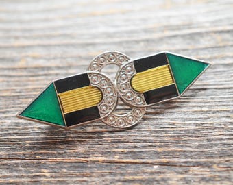 Könnte beinhalten: Eine silberne Brosche mit einem schwarzen, goldenen und grünen Emaille-Design. Die Brosche ist in Form von zwei ineinandergreifenden Pfeilen gestaltet.