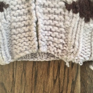 May include: A close-up of a gray knitted sweater with a brown and green pattern. The sweater has a zipper and is showing signs of wear.