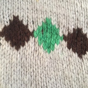 May include: Close-up of a knitted fabric with a brown and green diamond pattern. The fabric is a light gray color.
