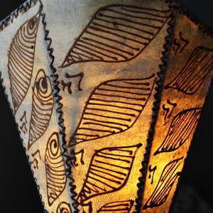 May include: A white lampshade with a brown, intricate pattern of leaves and lines. The lampshade is made of a textured material and has a fringed edge.