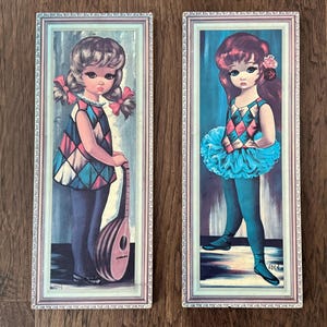 May include: Two framed vintage prints featuring large-eyed girls. One girl has pigtails and holds a lute, wearing a harlequin-patterned top. The other girl has red hair, a blue tutu, and a flower, also wearing a harlequin top. Both are in ornate frames.