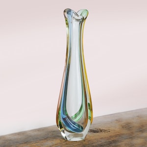 May include: A tall, clear glass vase with a flared top and a slender body. The vase has a swirl of colors, including blue, green, and yellow. The vase is sitting on a wooden surface.