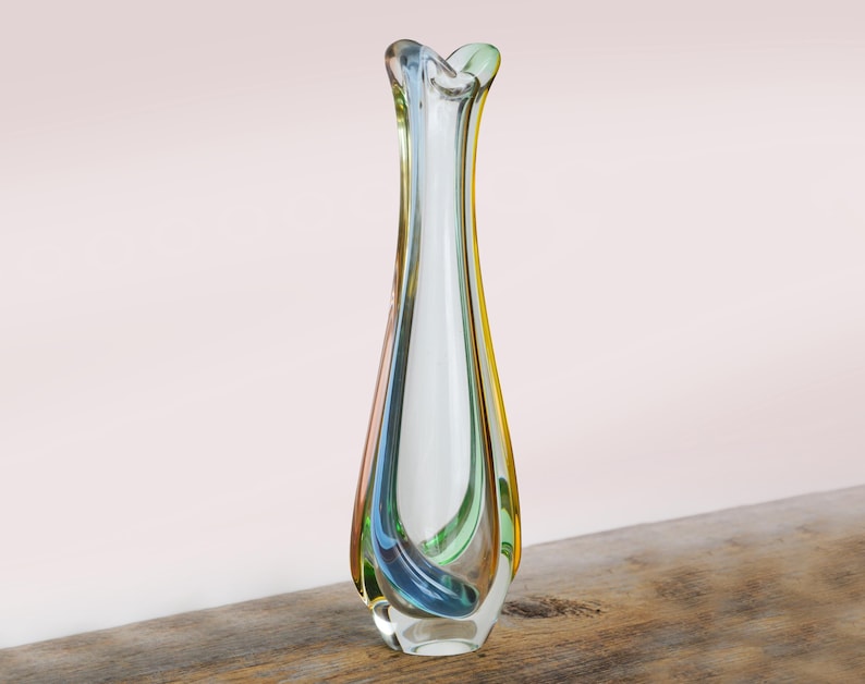 May include: A tall, clear glass vase with a flared top and a slender body. The vase has a swirl of colors, including blue, green, and yellow. The vase is sitting on a wooden surface.
