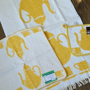 May include: Set of three white kitchen towels with yellow teapots and cups printed on them. The towels have a fringe border. The largest towel has the text "Cannon Kitchen Fashions" printed on it.