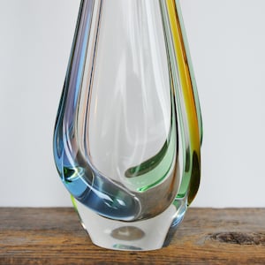 May include: A clear glass vase with a teardrop shape. The vase has streaks of blue, green, and yellow. The vase is sitting on a wooden surface.