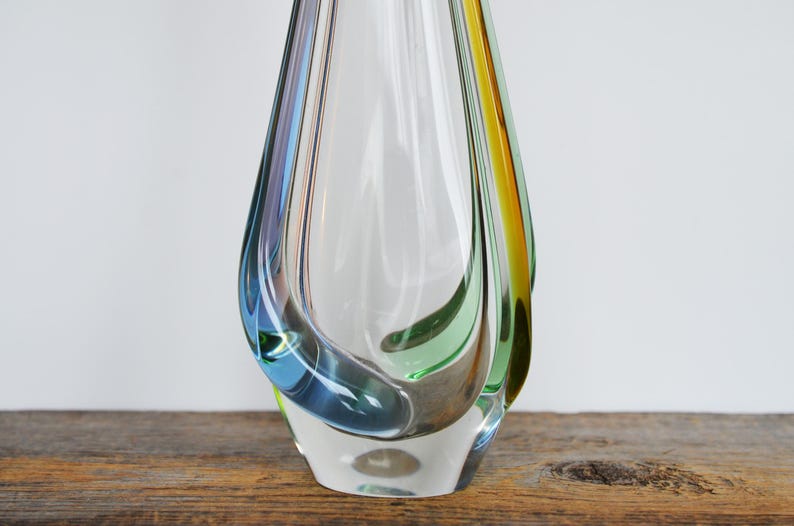 May include: A clear glass vase with a teardrop shape. The vase has streaks of blue, green, and yellow. The vase is sitting on a wooden surface.