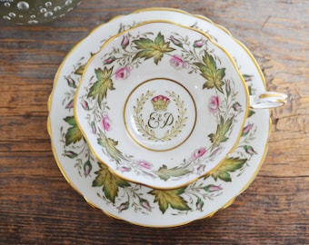 Könnte beinhalten: Ein Vintage-Tee-Set. Die weiße Porzellantasse und Untertasse sind mit einem floralen Muster aus rosa Rosen und grünen Blättern mit Goldrand verziert. Die Tasse zeigt eine Krone und die Initialen "E & D".