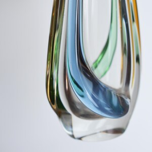May include: A clear glass vase with a unique, flowing design. The vase features streaks of blue, green, and amber colors that swirl within the transparent glass. The vase has a rounded shape with a narrow opening, suitable for decorative purposes.