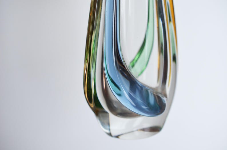 May include: A clear glass vase with a unique, flowing design. The vase features streaks of blue, green, and amber colors that swirl within the transparent glass. The vase has a rounded shape with a narrow opening, suitable for decorative purposes.