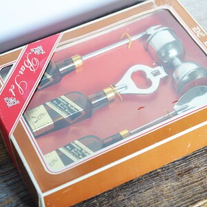 May include: A brown box with a red and white label that says "Bar Set" and "De Luxe". The box contains a set of four silver bar tools: a bottle opener, a jigger, a spoon, and a bottle stopper.