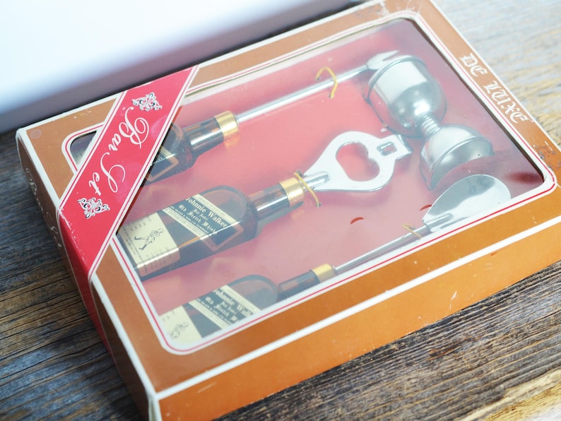 May include: A brown box with a red and white label that says "Bar Set" and "De Luxe". The box contains a set of four silver bar tools: a bottle opener, a jigger, a spoon, and a bottle stopper.