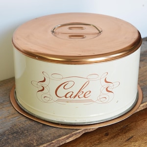 May include: A vintage cake carrier with a copper-colored lid and rim, and a cream-colored body. The word "Cake" is written in a decorative font on the front. The carrier sits on a wooden surface.
