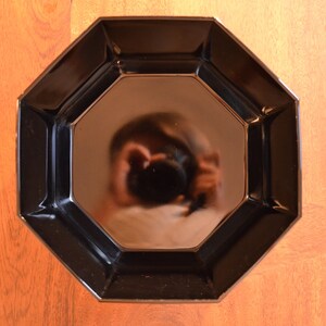 Pode incluir: Um prato octogonal preto com o reflexo de uma pessoa segurando uma c&acirc;mera no centro.