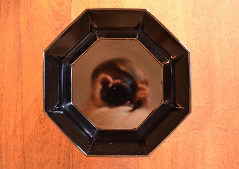 Pode incluir: Um prato octogonal preto com o reflexo de uma pessoa segurando uma c&acirc;mera no centro.