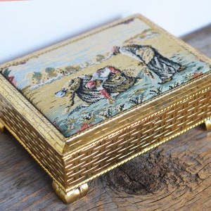 May include: A gold-colored metal box with a woven pattern on the sides and a tapestry lid featuring a colorful scene of people in a landscape.