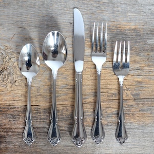 May include: A set of five silver-toned cutlery pieces, including a knife, two forks, and two spoons, arranged on a wooden surface.