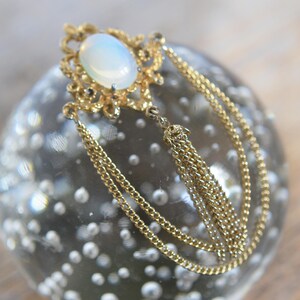 May include: A gold pendant with a white opal stone and a chain with a tassel. The pendant is on a clear glass surface with bubbles.