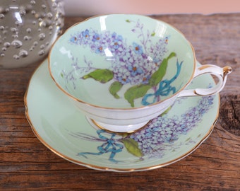 May include: A delicate teacup and saucer set with a light green background and a floral design featuring purple and blue flowers. The set has a gold trim around the edges.