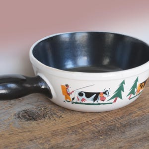 May include: A white ceramic fondue pot with a black interior and handle. The exterior features a painted scene with a cow, a person, and trees. The pot sits on a wooden surface.