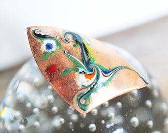 May include: A copper-toned, abstract enamel brooch with a swirling design. The brooch features a mix of colors, including blue, green, yellow, and red, creating a unique and artistic appearance. The brooch is a geometric shape.
