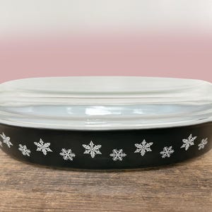 Puede incluir: Una fuente de horno ovalada de vidrio negro con un patrón repetitivo de copos de nieve blancos en los lados exteriores, cubierta con una tapa de vidrio transparente que tiene asas integradas en ambos extremos para un agarre seguro.
