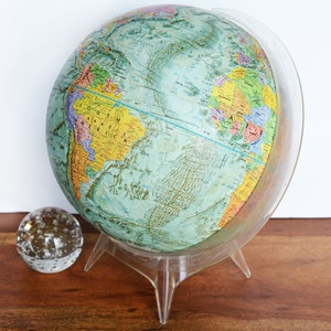 May include: A vintage globe with a blue and green map of the world. The globe is on a clear plastic stand with three legs. The globe is on a wooden surface.