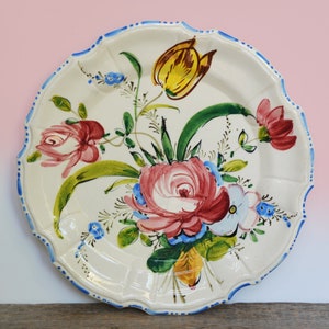 Puede incluir: Un plato de cerámica blanco con borde festoneado presenta un diseño floral pintado a mano. Las flores son rosas, rojas, amarillas, azules y verdes.