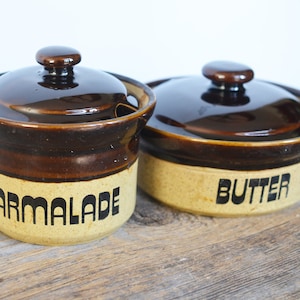 Op de afbeelding: Twee bruine keramische potten met deksels. De ene pot is geëtiketteerd met "MARMALADE" en de andere met "BUTTER".