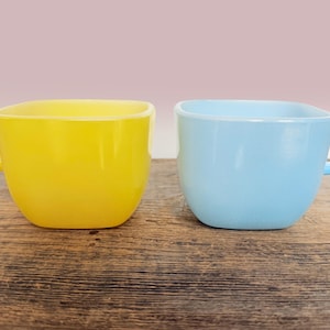 Pode incluir: Duas canecas de formato quadrado feitas de vidro liso e opaco. Uma caneca é de cor amarela sólida e brilhante com uma alça no lado esquerdo, e a outra caneca é de cor azul-celeste sólida e clara com uma alça no lado direito. Cantos arredondados.