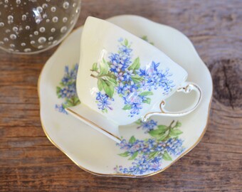 May include: A white teacup and saucer with a floral pattern of blue and green flowers. The cup and saucer have a gold trim.