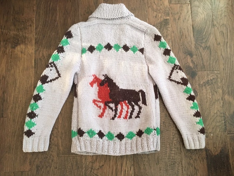 May include: A light purple knitted cardigan with a brown and green geometric pattern and two red and brown horses on the back.