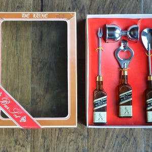 May include: A red and gold box with a clear window showing a bar set. The set includes three miniature bottles, a bottle opener, a jigger, and a cocktail spoon. The box says "De Luxe Bar Set" on the front.